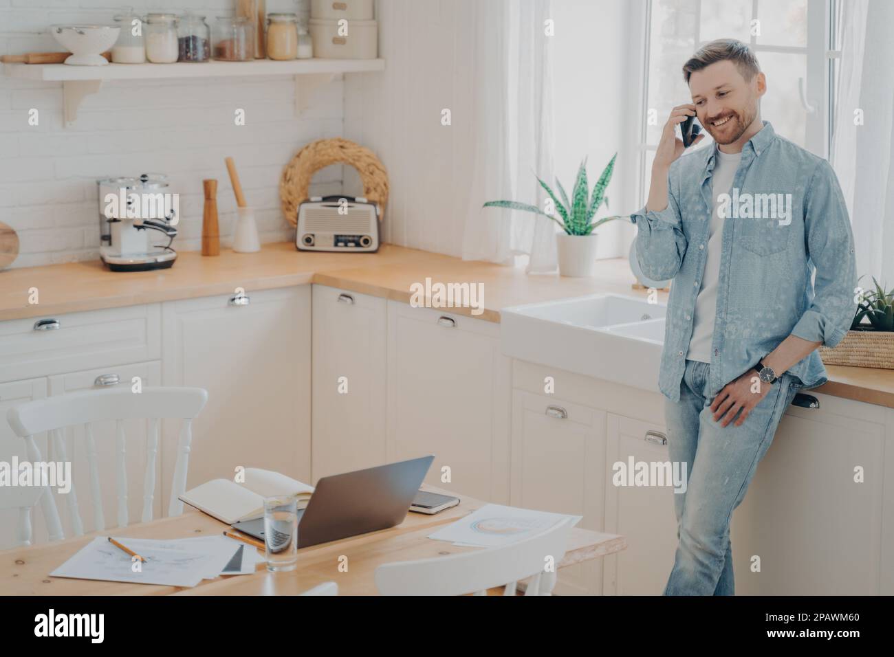 Ein glücklicher junger Kaukasier, der ein Handy in der Hand hält, während er sich mit dem Kunden unterhält, in der modernen Küche steht und ein gutes Geschäftsgespräch genießt Stockfoto