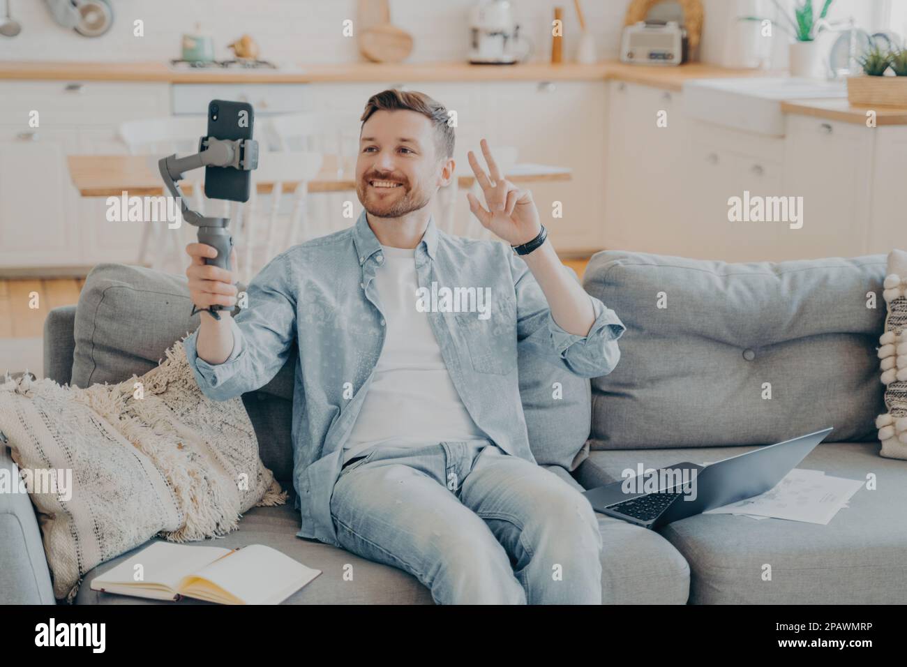 Positiver junger Mann mit Bart, der während eines Videogesprächs ein Friedenszeichen zeigt, sitzt auf einer bequemen Couch mit Laptop und Papieren, offenes Notizbuch neben ihm, verschwommen Stockfoto