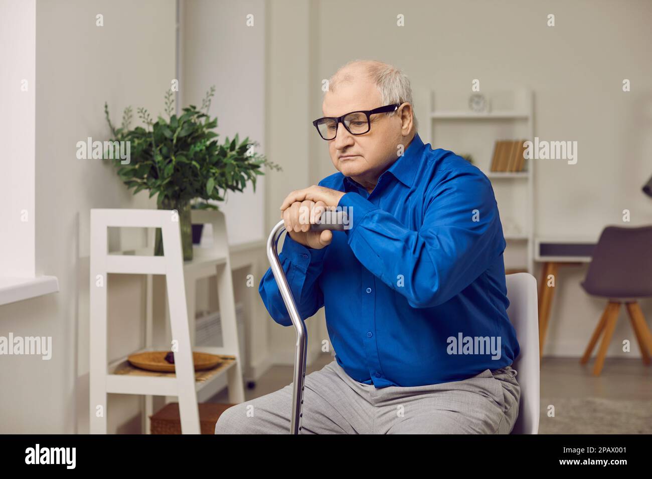 Seniorenmann, der zu Hause in der Rehabilitation sitzt, mit seiner Krücke und traurig mit Brille aussieht. Stockfoto