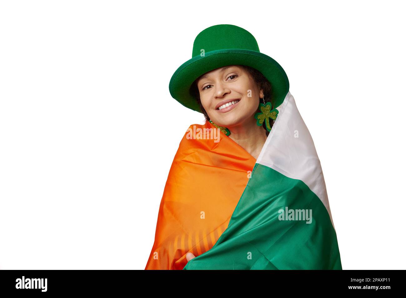 Lächelnde Brünette, eingewickelt in irische Flagge, trägt einen grünen Hut mit Kleeblättern, lächelt vor die Kamera auf weißem Hintergrund Stockfoto