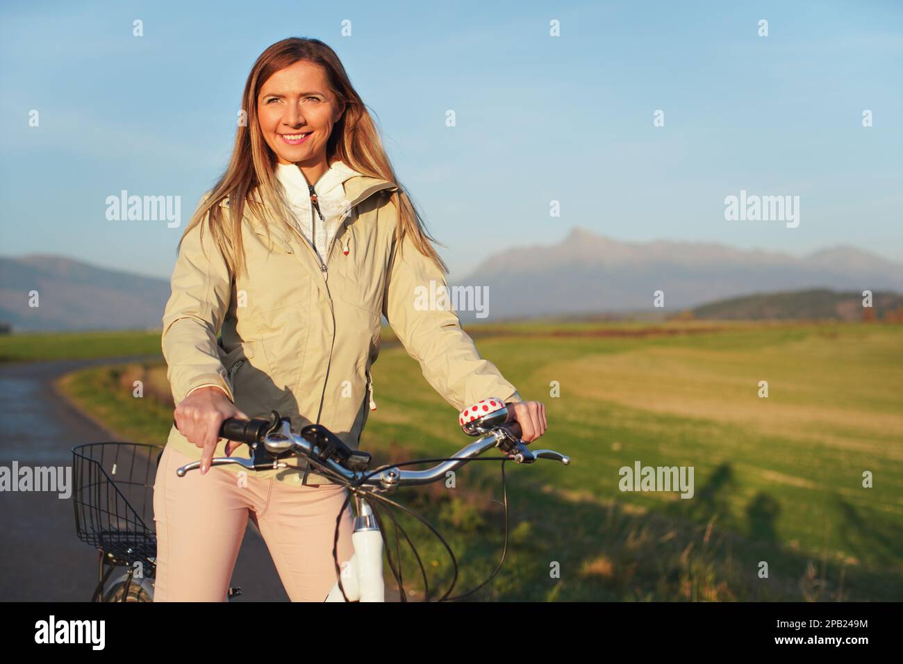 Junge Frau in Jacke steht mit dem Fahrrad auf Asphalt Landstraße, Nachmittag Sonne scheint auf Feldern Wiesen und Berge Hintergrund Stockfoto