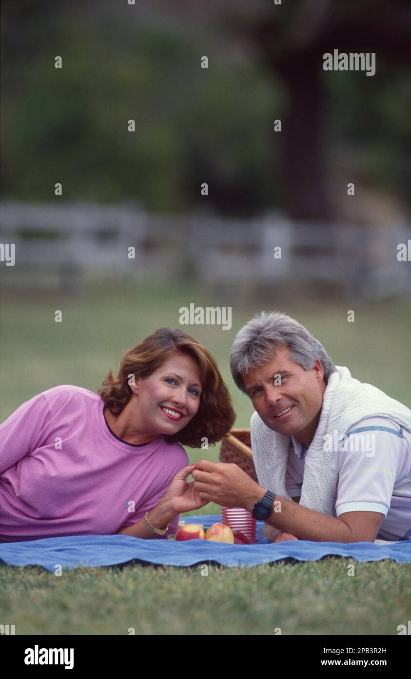 Ein Paar mittleren Alters an einem wunderschönen Tag im Park, das auf einer Decke liegt, sich gegenüberliegt und beim Picknick Händchen hält Stockfoto