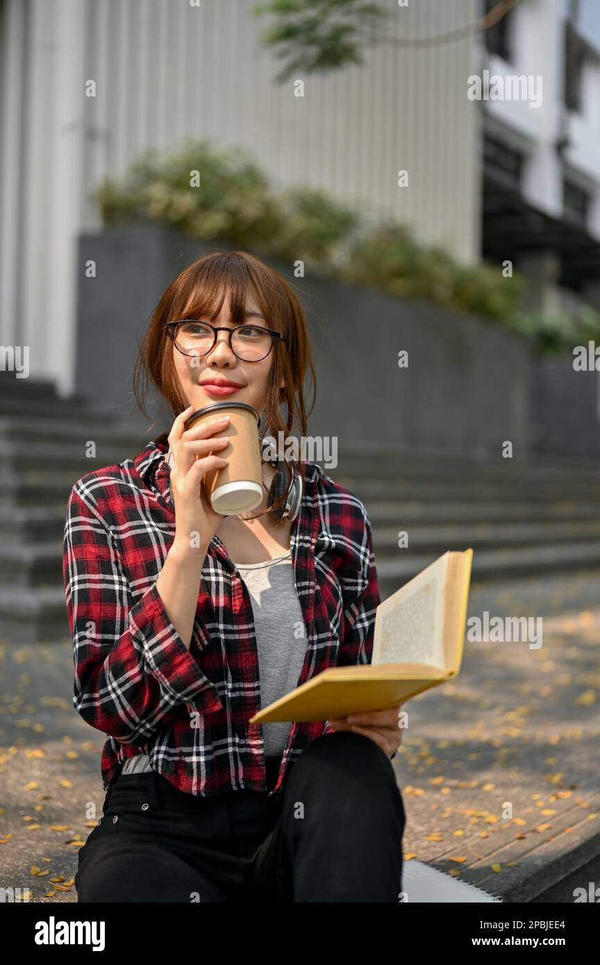 Porträt, charmante junge asiatische Studentin in lässiger Kleidung, trinkt Kaffee, während sie auf der Treppe vor dem Campus-Gebäude Buch liest Stockfoto