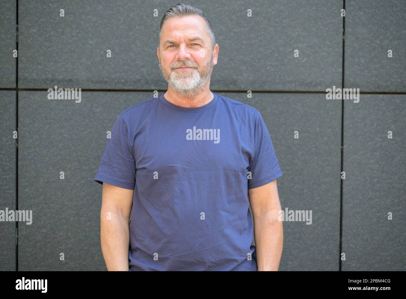 Ein bärtiger Mann mittleren Alters in einem Oberkörperporträt, der in einem Sommer-T-Shirt vor einer grauen Wand steht und in die Kamera lächelt Stockfoto