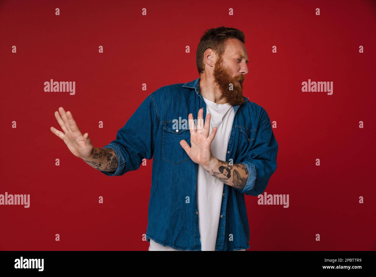 Ein erwachsener, mit Bart tätowierter, gutaussehender rothaariger Mann, der eine Abstoßungsgeste mit geschlossenen Augen macht und seinen Kopf wegdreht, während er über einem isolierten roten Backg steht Stockfoto