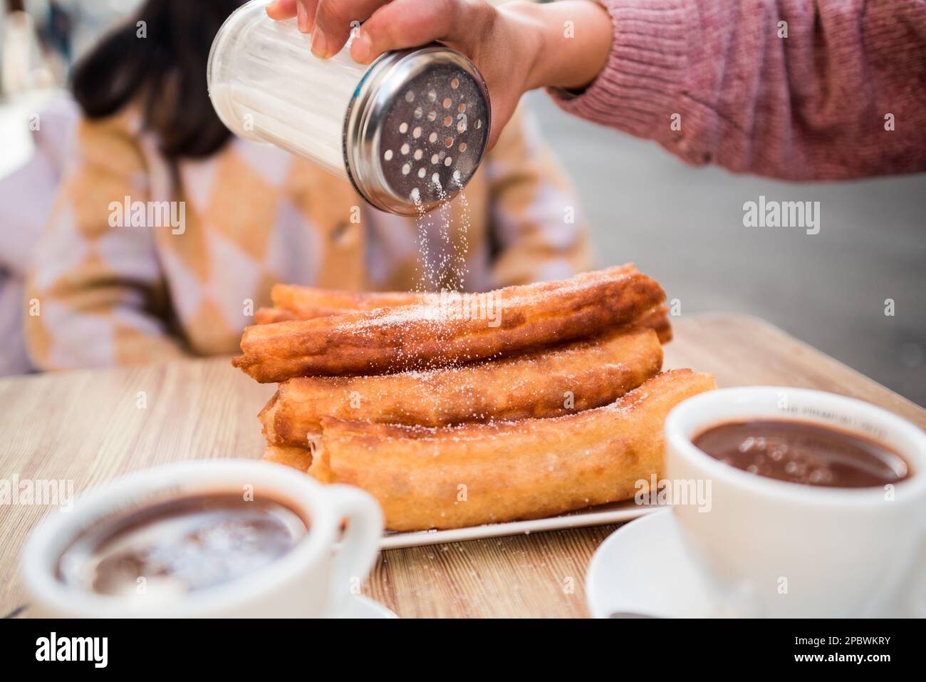 Die Hand einer Frau gießt Zucker auf die Churros, die auf dem Bartisch serviert werden. Stockfoto