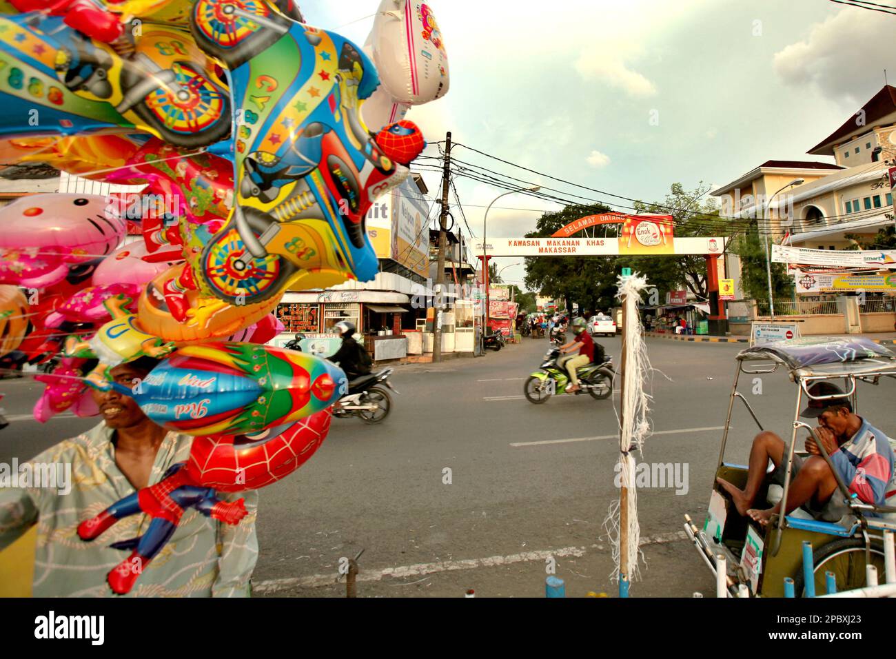 Ein mobiler Ballonverkäufer und Rikscha-Fahrer vor dem Hintergrund des Straßenverkehrs und einer Kreuzung der Penghibur Street, wo geschäftige wirtschaftliche Aktivitäten stattfinden, mit der Datu Musseng (Museng) Street, einem Zentrum der traditionellen Speisen, in der Küstenregion von Makassar City, South Sulawesi, Indonesien. Als eine der 30 Küstenstädte Indonesiens, die schätzungsweise vom steigenden Meeresspiegel betroffen sein wird, wurde Makassar von Forschern und Wissenschaftlern als Stadt mit erheblichen ökologischen und sozialen Problemen betrachtet, vor allem als Folge der geomorphologischen Bedingungen und der Landnutzungspolitik Stockfoto