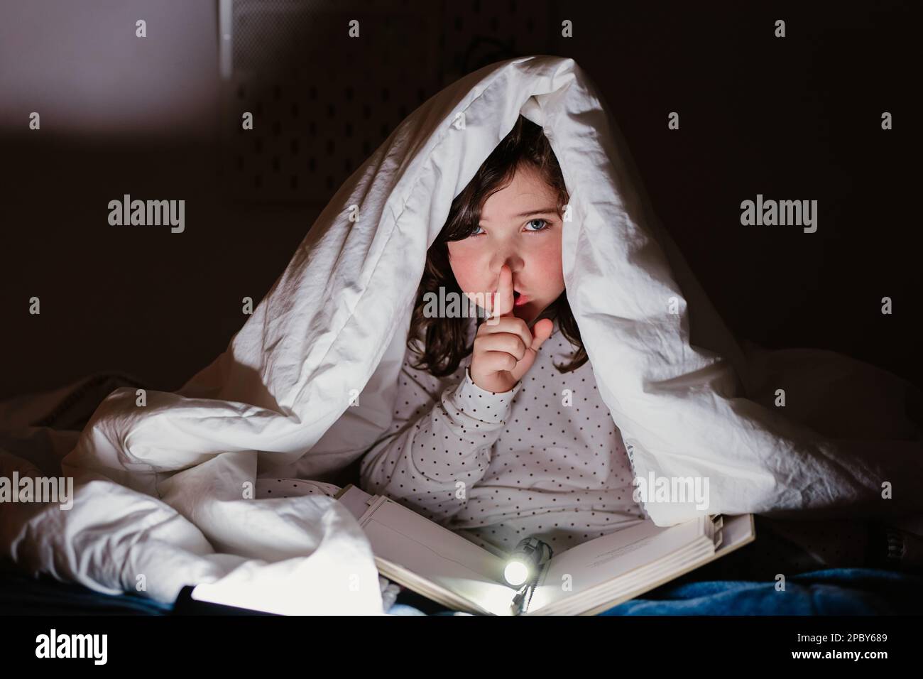 Fröhliches junges Mädchen im weißen Pyjama, das auf dem Bett sitzt und ein Buch mit Taschenlampe liest, während es sich unter der Decke versteckt und zeigt, wie es ist Stockfoto