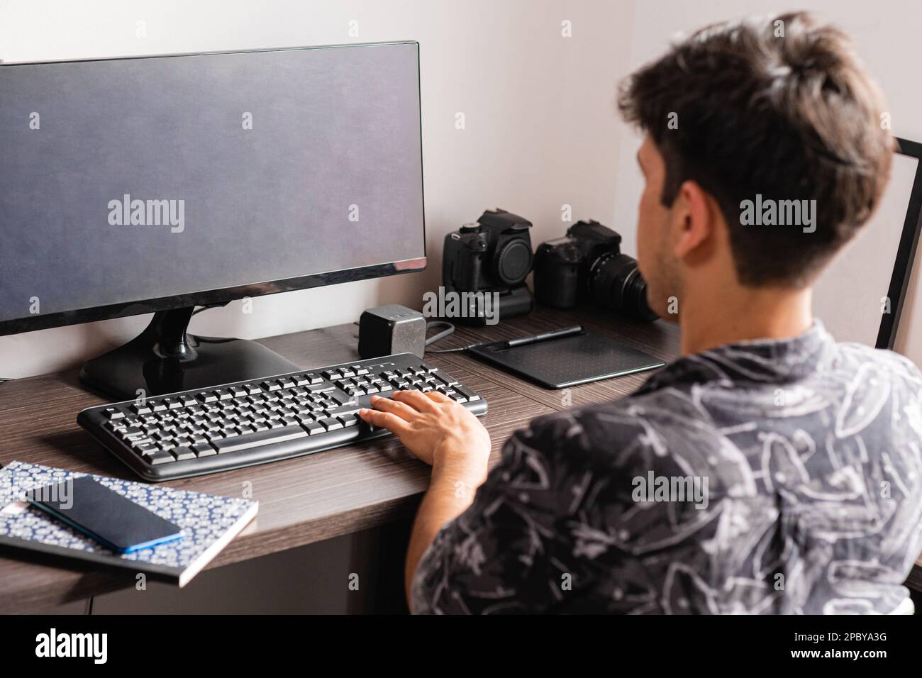 Rückansicht des ganzen Körpers eines nicht wiedererkennbaren männlichen Freiberuflers in schwarzem T-Shirt mit weißen Blumen, der auf einem bequemen Stuhl am Holzschreibtisch sitzt und Com benutzt Stockfoto