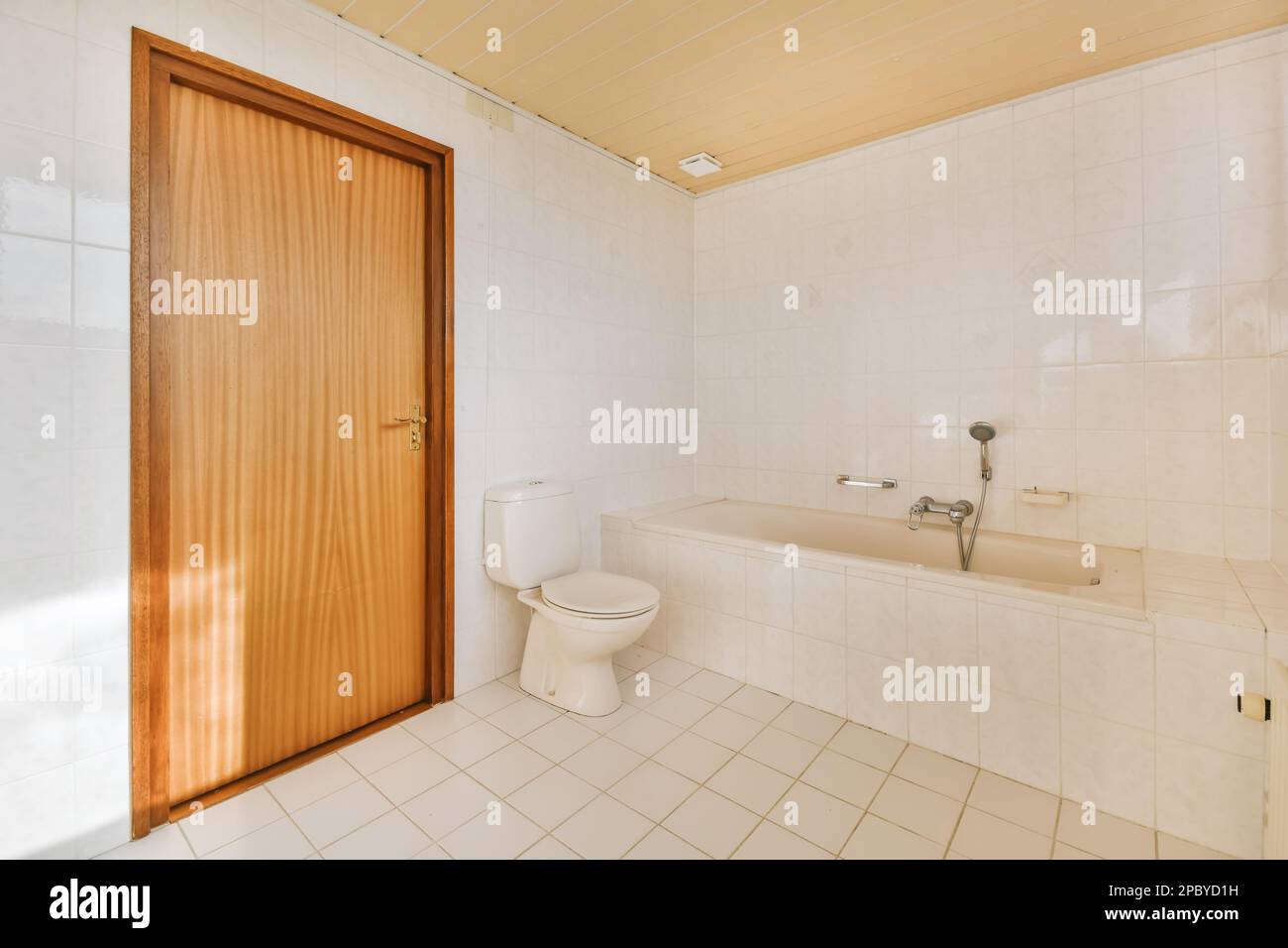 Helles Badezimmer mit weißer Toilette und Badewanne neben Holztür an gefliesten Wänden des Apartments Stockfoto