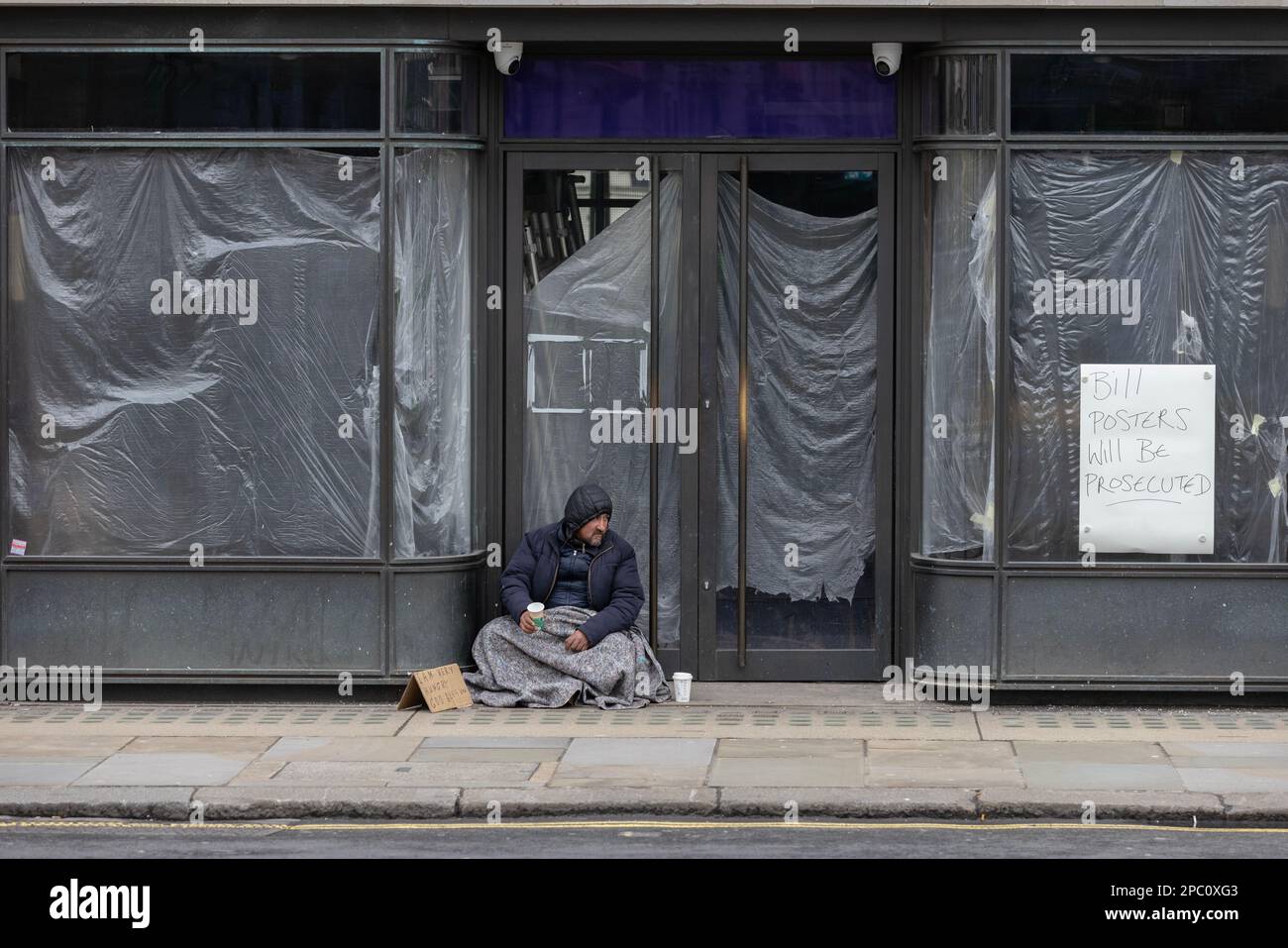 Obdachloser sitzt in einer leeren, verkleideten Ladenfront, die geschlossen wurde, weil die britische Einzelhandelswirtschaft unter hoher Inflation leidet, London, England. Stockfoto
