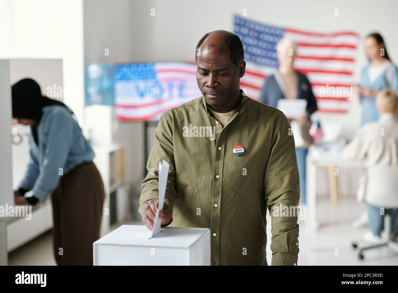 Reifer afroamerikanischer Mann, der an Präsidentschaftswahlen teilnimmt, während er an der Wahlurne steht und ein Dokument mit seiner Wahl dort ablegt Stockfoto