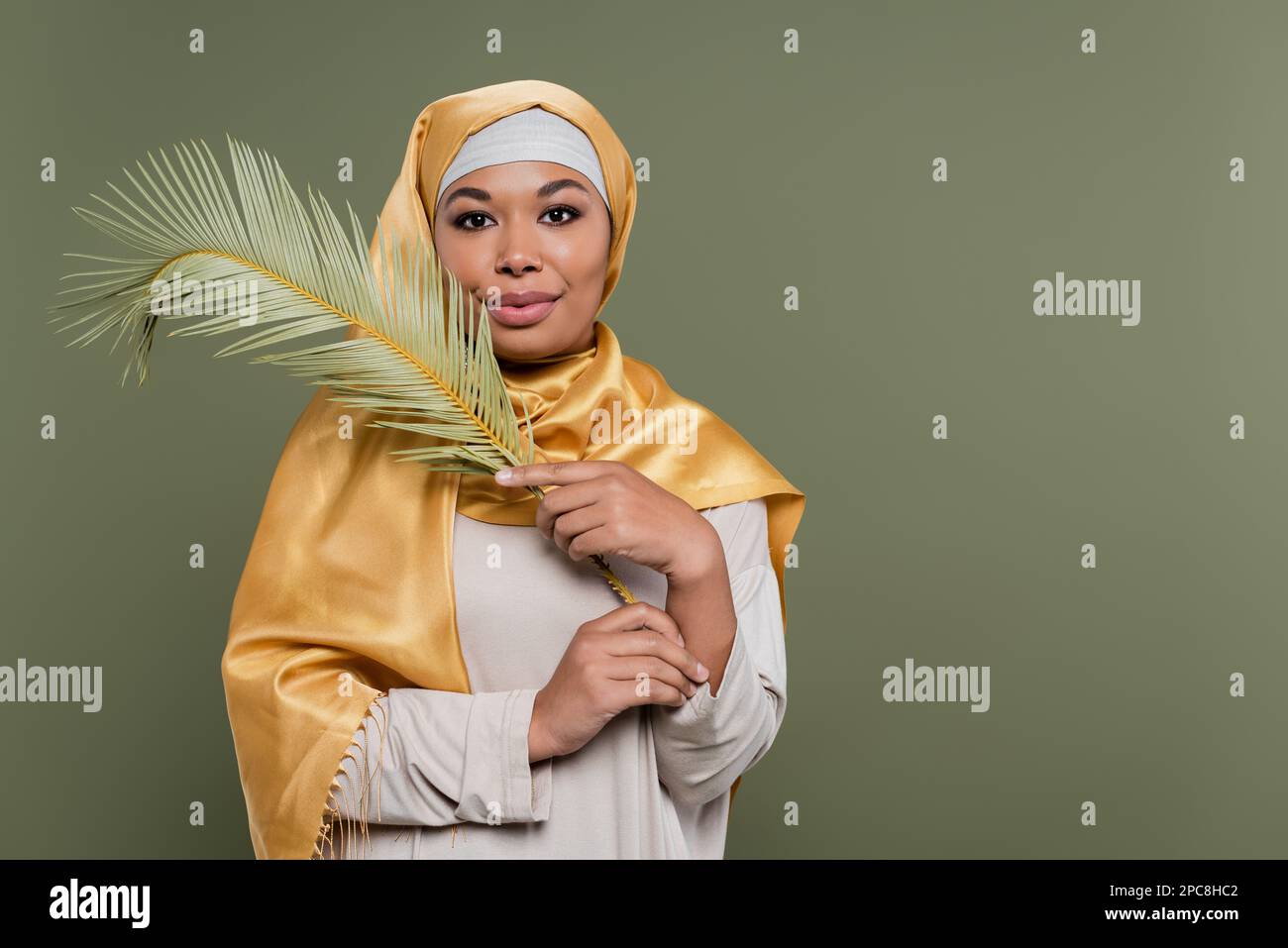 Glückliche Multirassenfrau mit Make-up, trägt gelben Satin-Hijab und hält exotische Blätter isoliert auf grünem Stockbild Stockfoto