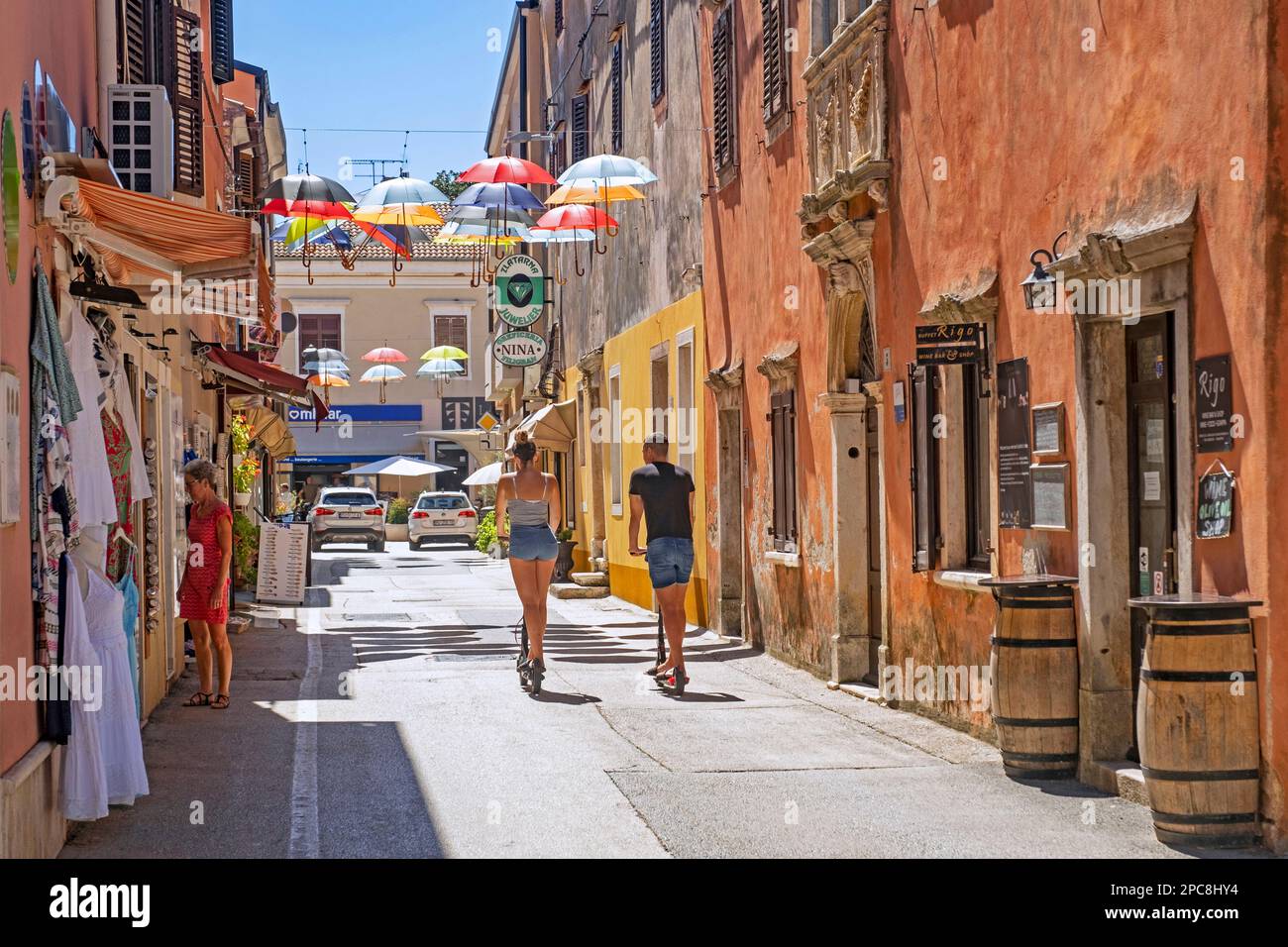 Touristen auf E-Scootern fahren durch die Einkaufsstraße mit bunten Sonnenschirmen im historischen Stadtzentrum von Novigrad, Istrien, Kroatien Stockfoto