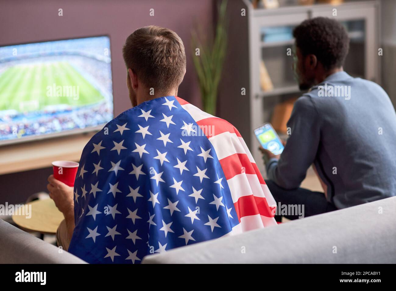 Rückansicht von zwei Männern, die ein Fußballspiel im Fernsehen sehen und die amerikanische Flagge tragen Stockfoto