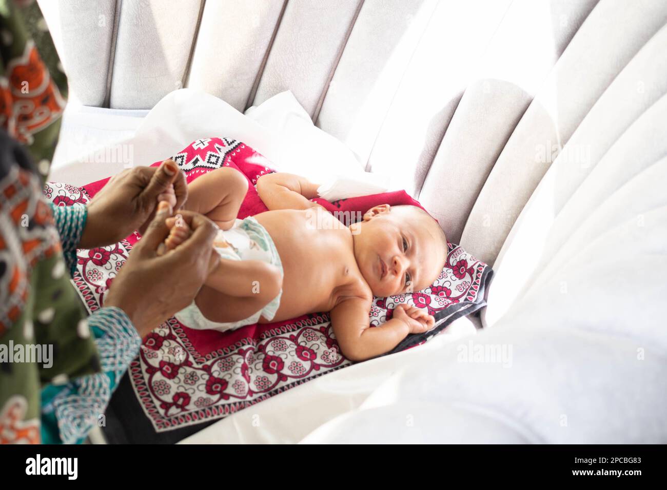 Phototherapie. Eine Frau mit einem Neugeborenen macht leichte Übungen mit Bewegungen der unteren Gliedmaßen, während das Baby natürliches Sonnenlicht durch das Fenster bekommt Stockfoto
