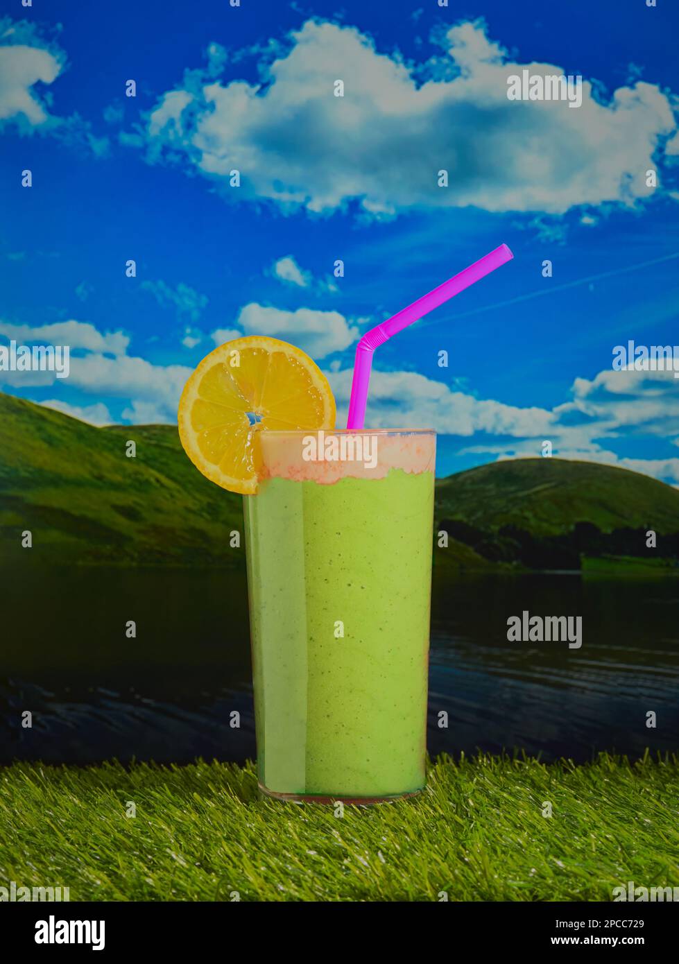 Lebhafter grüner Smoothie garniert mit Creme und einer Zitronenscheibe, aufgenommen auf einer Außenlandschaft mit Hügeln und blauem Himmel mit flauschigen Wolken. Stockfoto