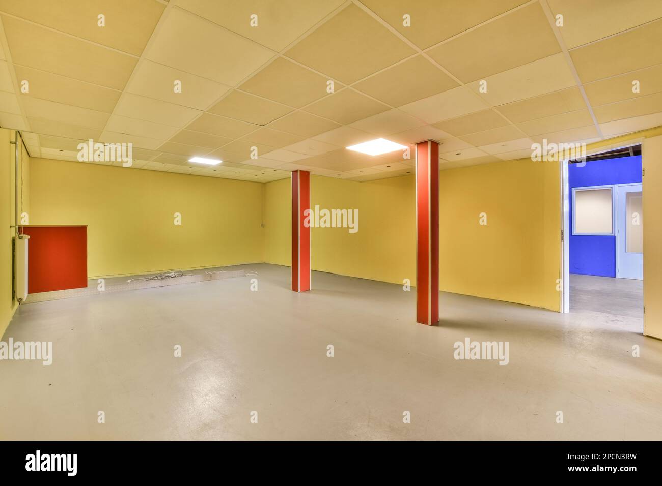 Ein leerer Raum mit gelben Wänden und weißen Deckenfliesen auf dem Boden, es gibt eine rote Tür in der Ecke Stockfoto