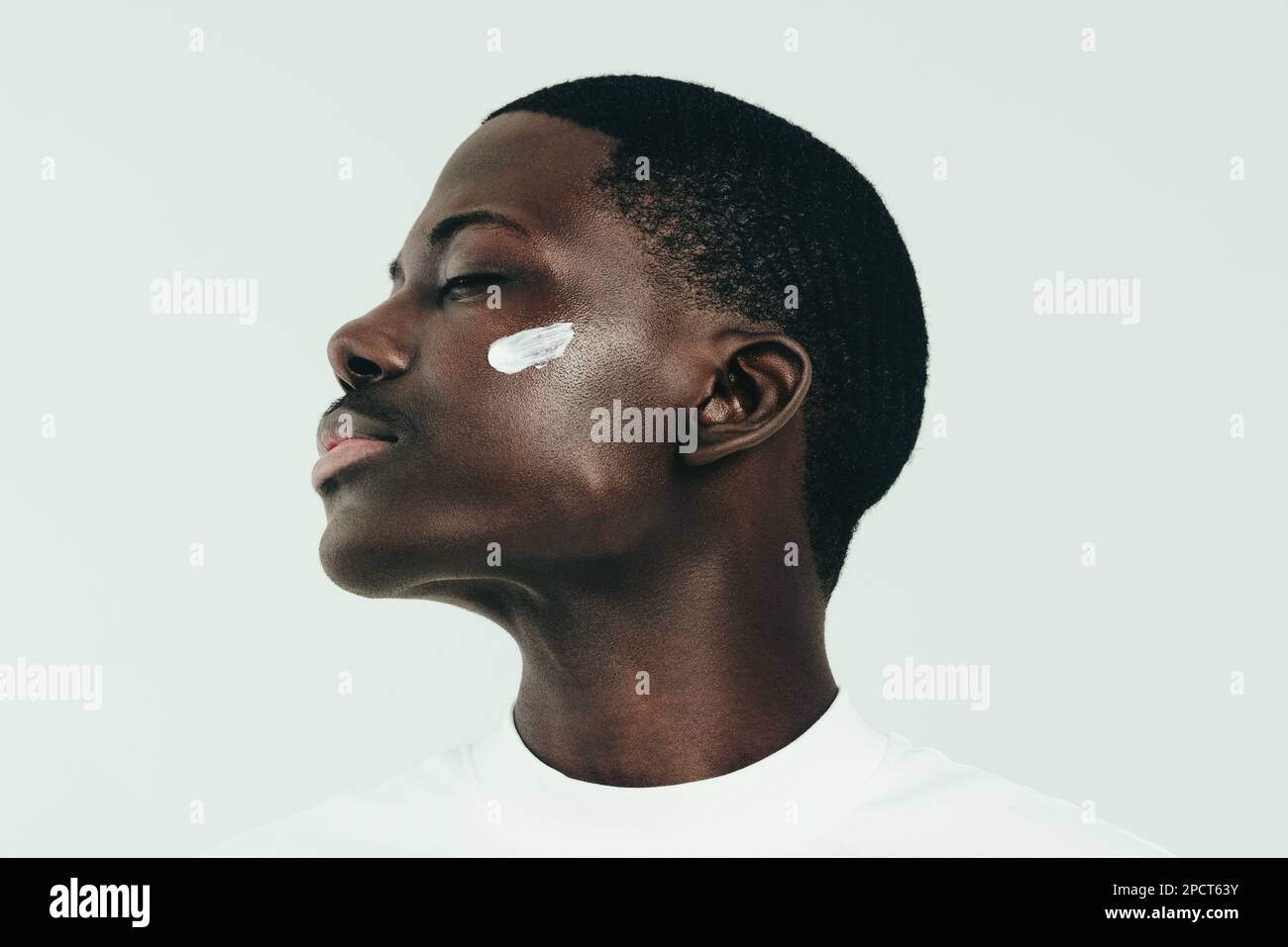 Ein junger schwarzer Mann, der sich um seine Haut kümmert und pflegende Gesichtscreme aufträgt, um sie feuchtigkeitsspendend und strahlend zu halten. Mit geschlossenen Augen schmeckt er das "R" Stockfoto