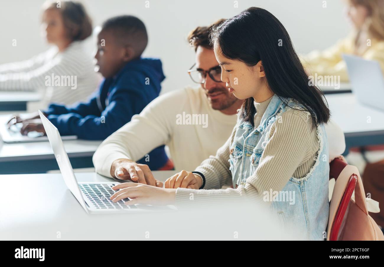 In einem Klassenzimmer voller Kinder hilft ein männlicher Lehrer einem jungen Schüler bei der Programmierung als Teil einer Informatikstunde. Bereitstellung von Lehrkräften für digitale Kompetenz Stockfoto