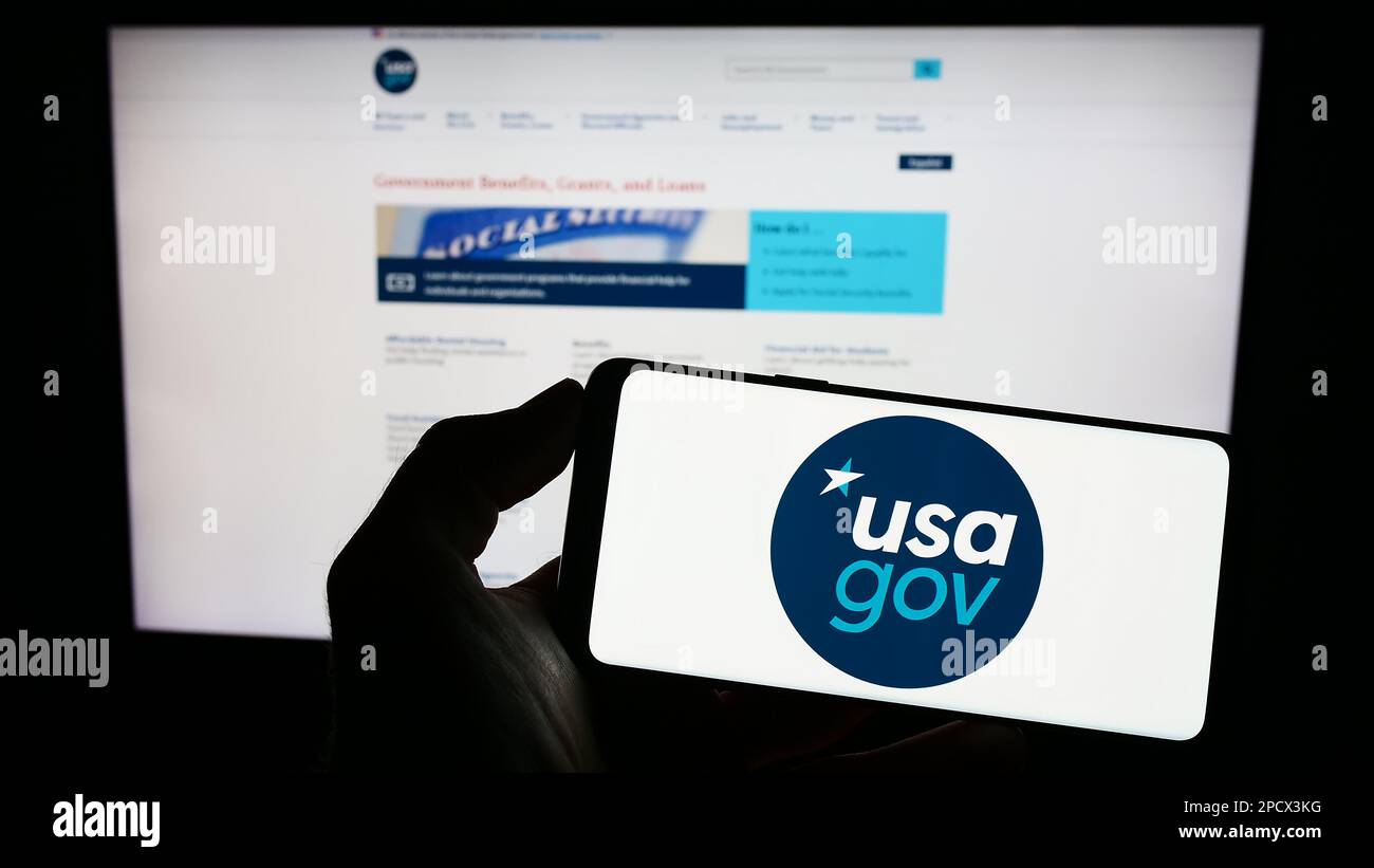 Person, die ein Mobiltelefon mit dem Logo der Website der US-Bundesregierung USA.gov auf dem Bildschirm vor der Webseite hält. Konzentrieren Sie sich auf das Display des Telefons. Stockfoto