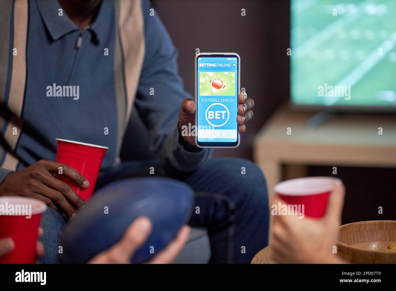 Nahaufnahme eines Mannes, der sein Smartphone mit der App für Sportwetten in die Kamera hält, während er sich zu Hause ein Fußballspiel ansieht, Kopierbereich Stockfoto