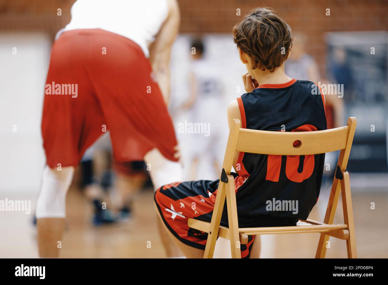 Junger Basketballspieler, Der Basketball-Übungsübungen Anschaut. Ein Junge sitzt auf einem Holzstuhl und ruht sich während des Basketballtrainings aus. Basketball-Camp Stockfoto