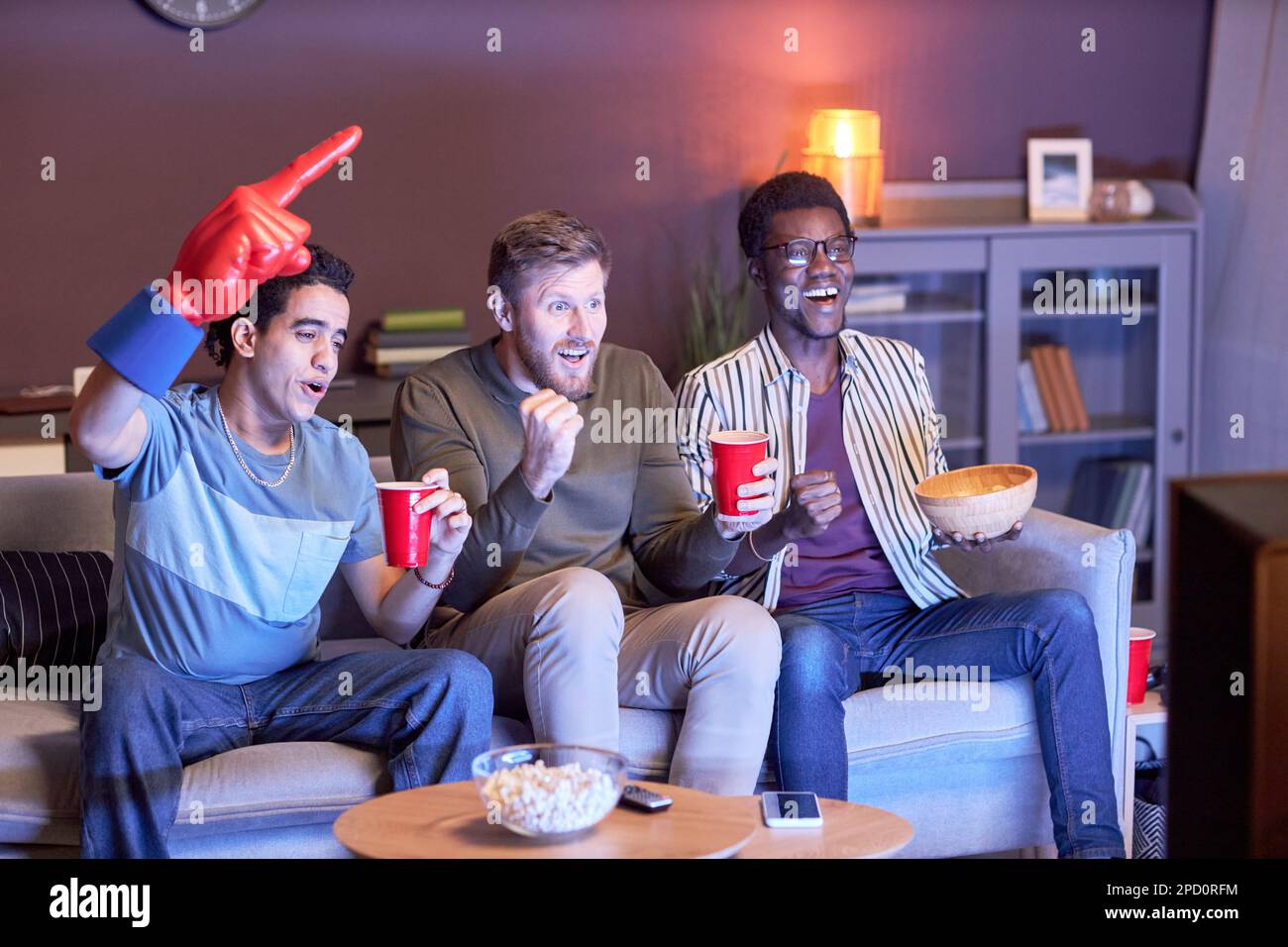 Eine Gruppe von emotionalen Sportfans, die zu Hause ein Spiel anschauen und von blauem Licht angestrahlt werden Stockfoto