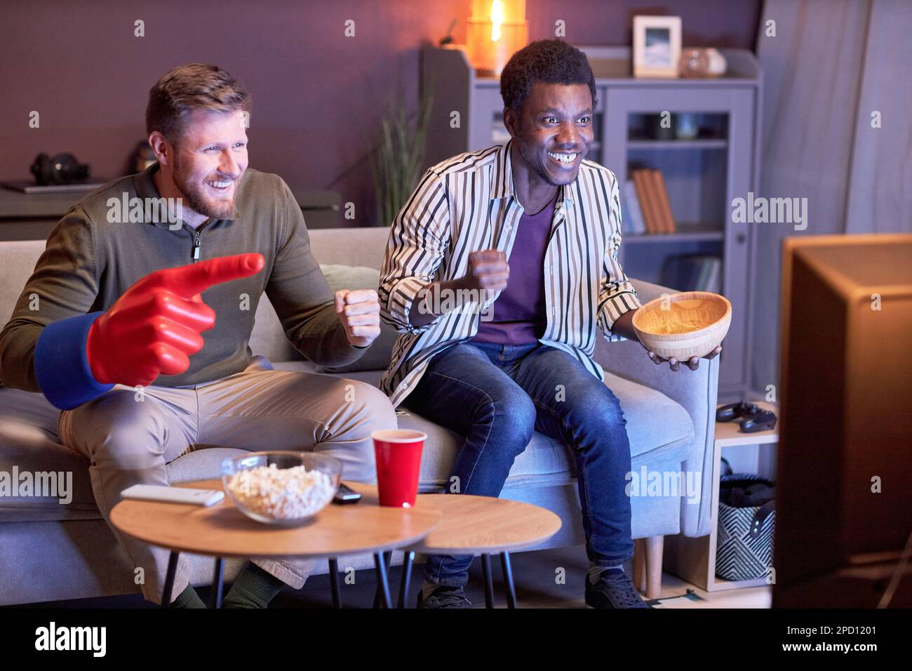 Porträt zweier emotionaler Sportfans, die in blauem Licht Fernsehen und Snacks essen Stockfoto