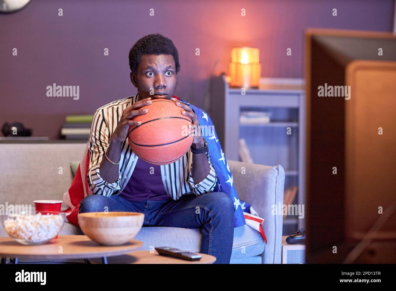 Porträt eines jungen Schwarzen als Sportfan, der zu Hause ein Basketballspiel im Fernsehen sieht und die Flagge der USA in blauem Licht trägt Stockfoto