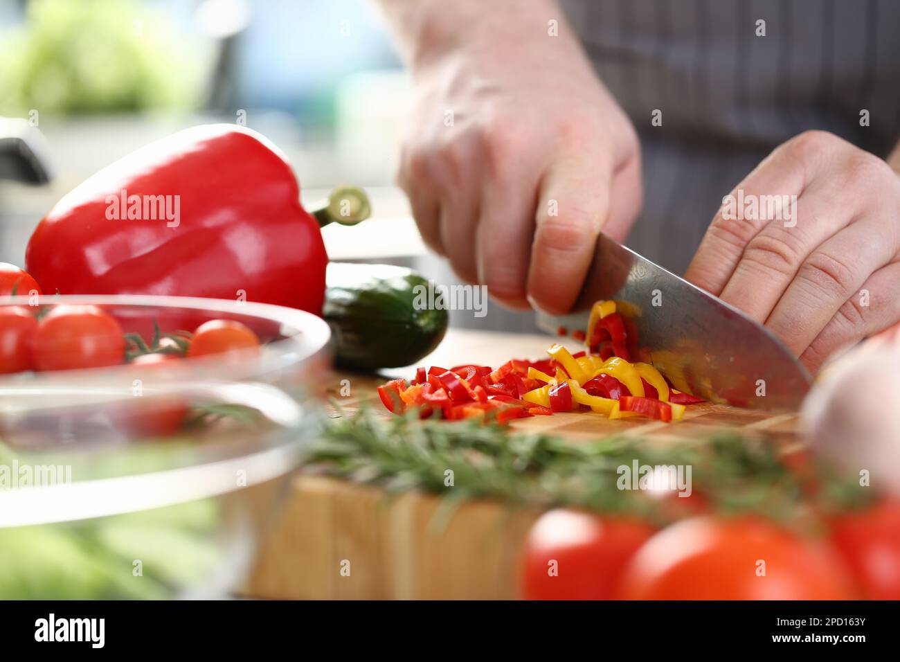 Die Hände des Kochs schneiden gelbe und rote Paprika auf dem Schneidebrett zusammen mit anderem Gemüse auf dem Tisch in der Küche Stockfoto