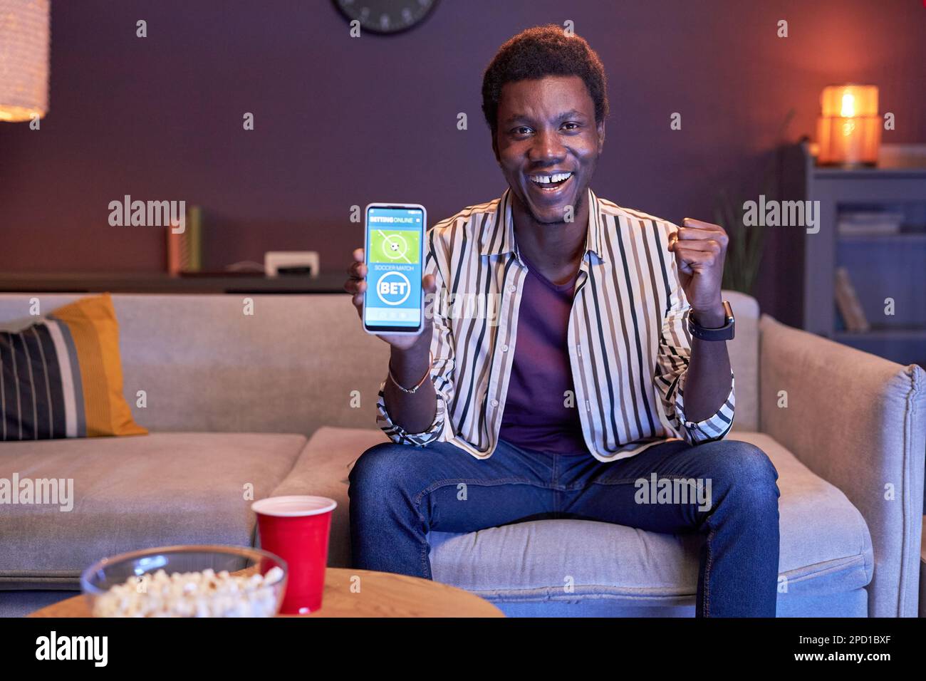 Porträt eines aufgeregten Schwarzen, der sich zu Hause ein Sportereignis im Fernsehen ansieht und die Online-Wett-App in blauem Licht und im Kopierbereich nutzt Stockfoto