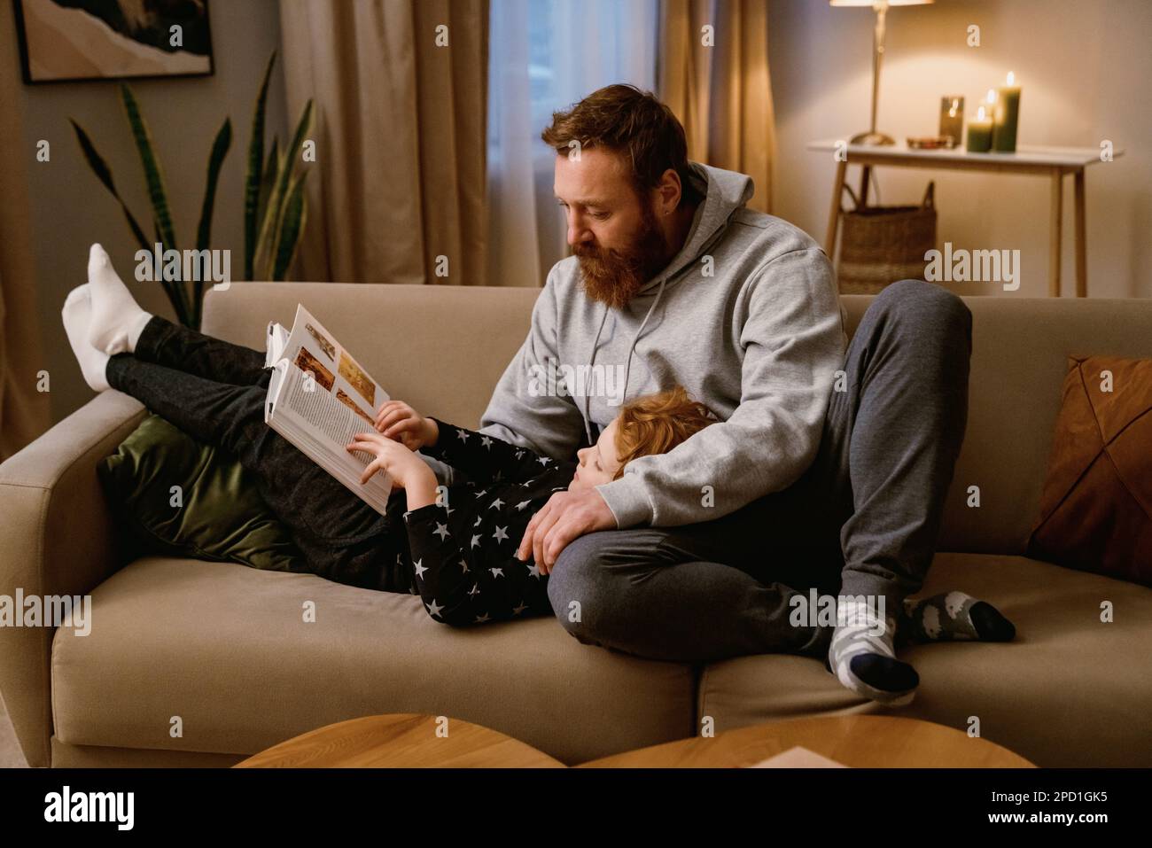 Vater und kleiner Sohn lesen Buch, während sie sich zusammen auf dem Sofa in einem gemütlichen Wohnzimmer zu Hause ausruhen Stockfoto