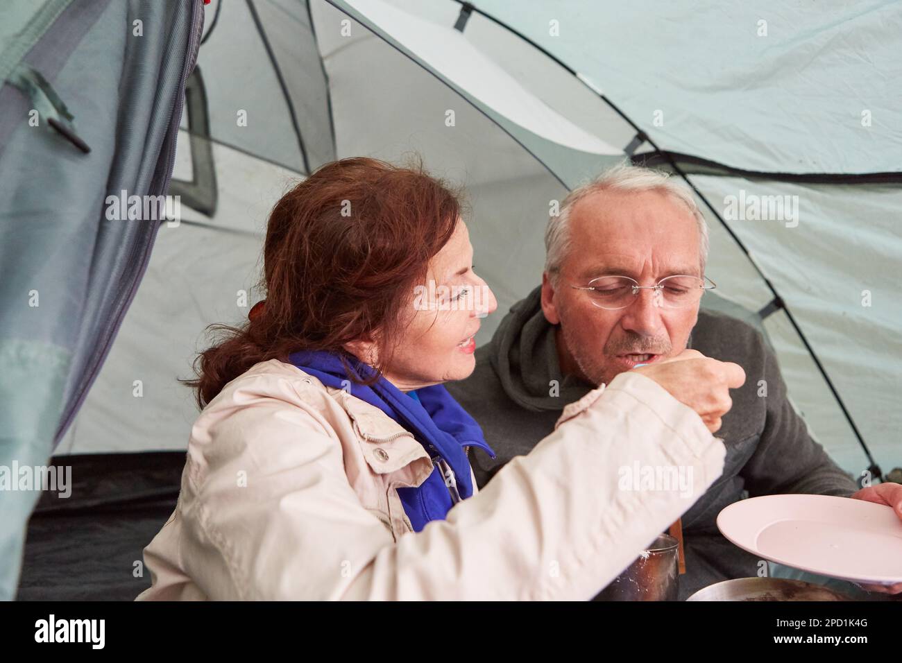 Ältere Frau füttert Mann mit Teller im Zelt während Camping im Urlaub Stockfoto