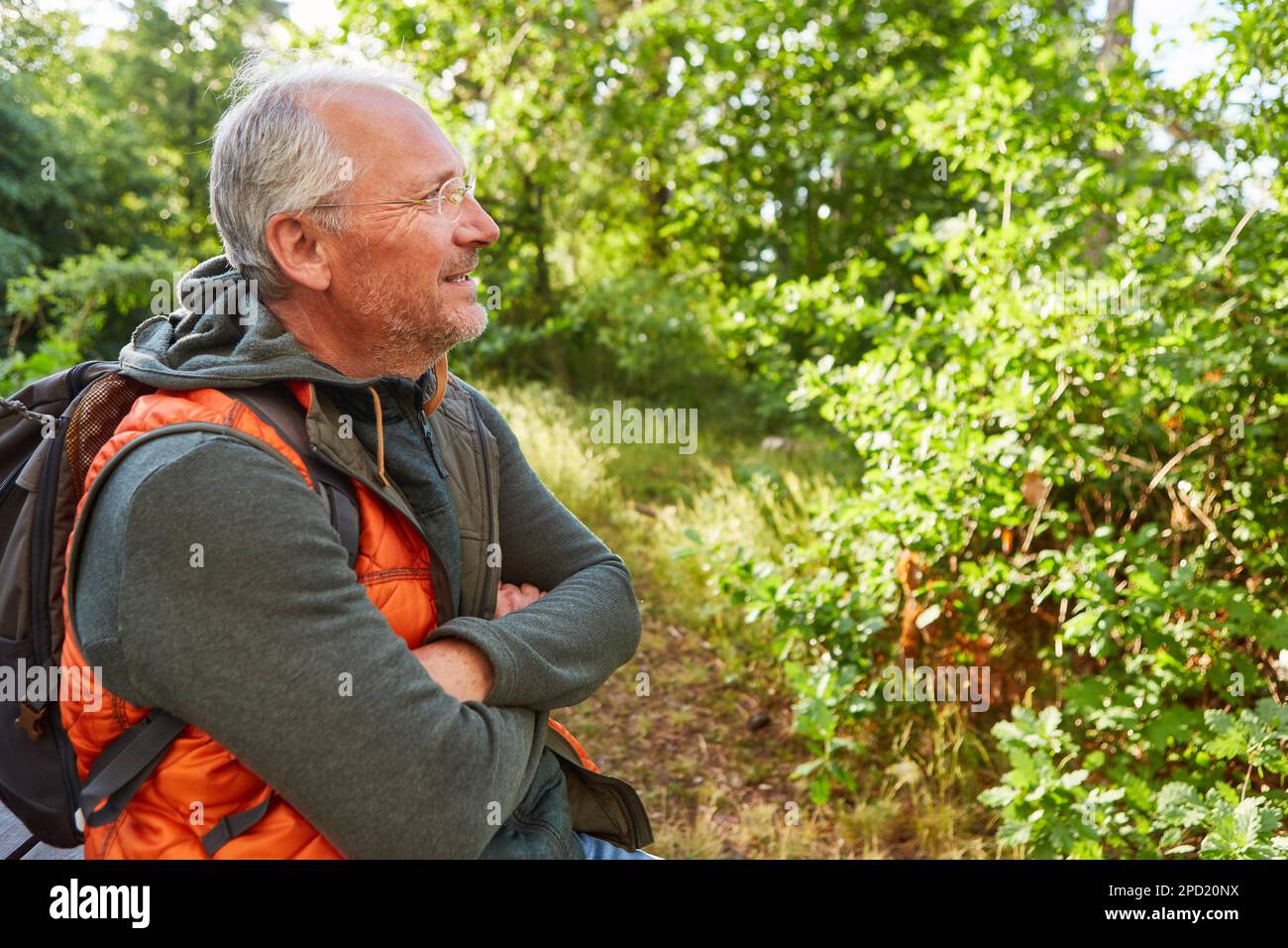 Lächelnder Seniorenmann mit Rucksack, der während der Sommerferien im Wald mit gekreuzten Armen stand Stockfoto