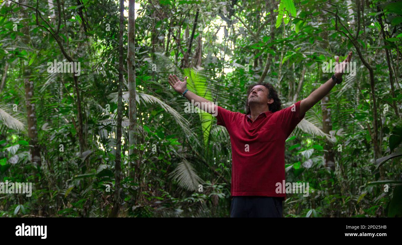 Ein junger Tourist mit rotem T-Shirt hebt die Arme und schaut an einem bewölkten Tag mitten in einem amazonischen Wald zum Himmel Stockfoto