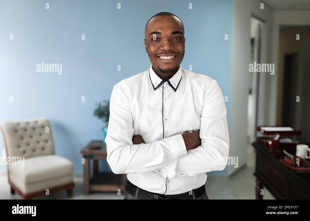 Ein fröhlicher, afrikanischer Mann steht in einem Innenbüro mit verschränkten Armen und einem lächelnden Gesicht, während er direkt in die Kamera schaut. Stockfoto