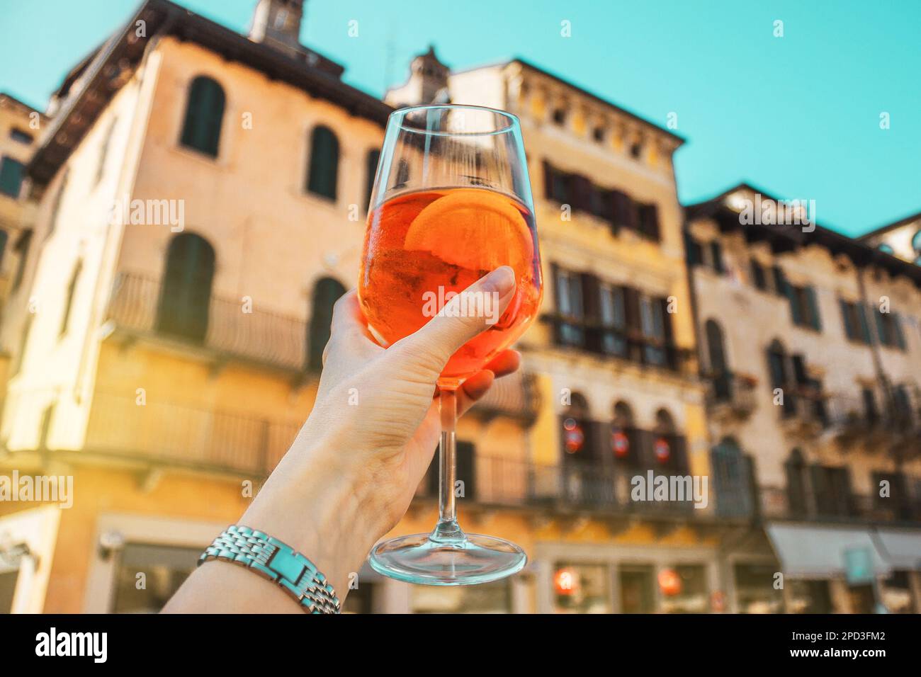 Weibliche Hand mit einem Glas orangefarbenem Cocktail Spritz in der Nähe von alten Gebäuden. Sonniger Tag in Verona, Italien Stockfoto