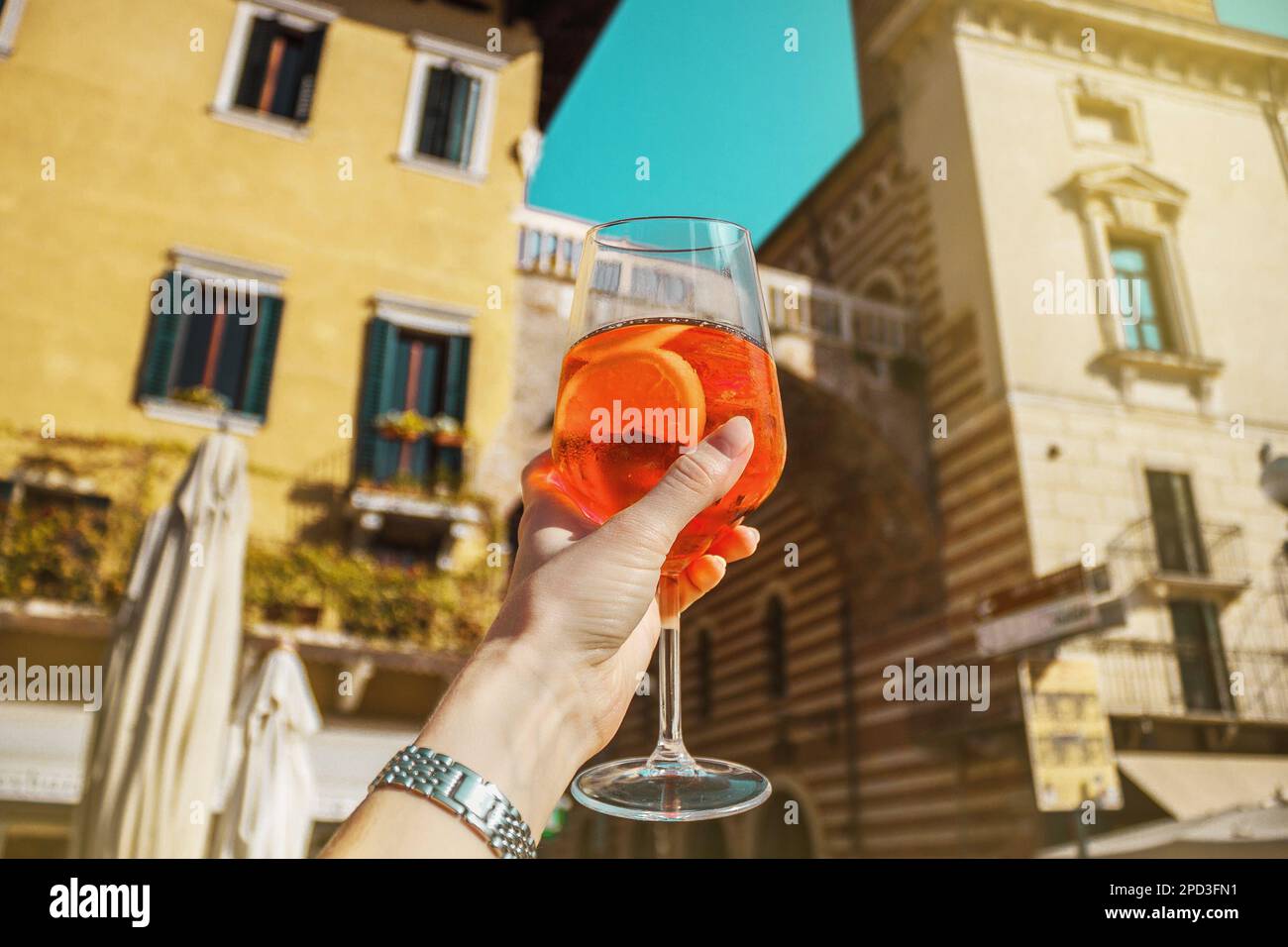 Weibliche Hand mit einem Glas orangefarbenem Cocktail Spritz in der Nähe von alten Gebäuden. Sonniger Tag in Verona, Italien Stockfoto