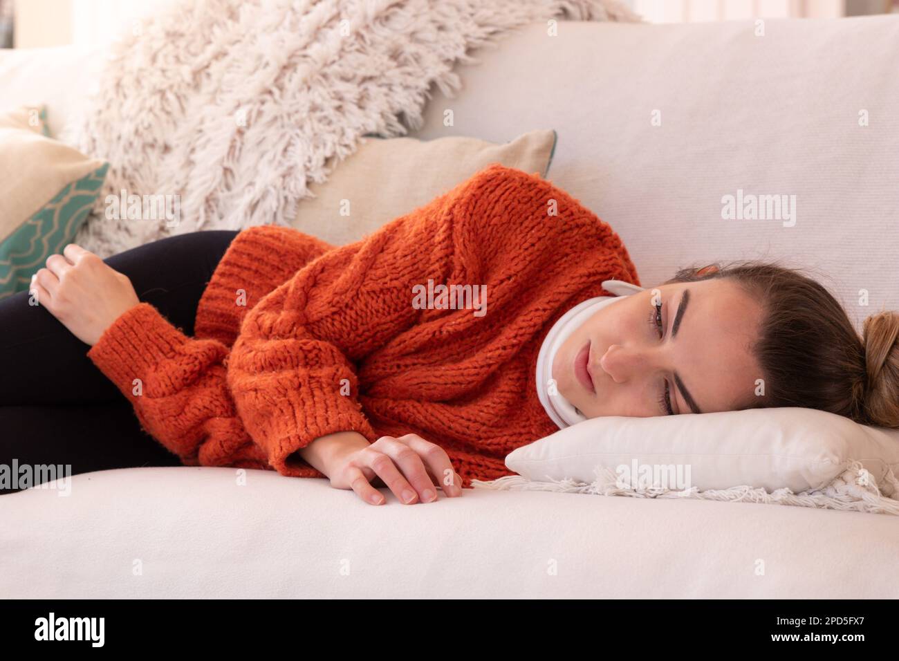Müde junge Frau in orangefarbener warmer Kleidung mit orthopädischem Kragen, die sich nach einer Zervixverletzung im Wohnzimmer zu Hause auf der Couch ruht Stockfoto