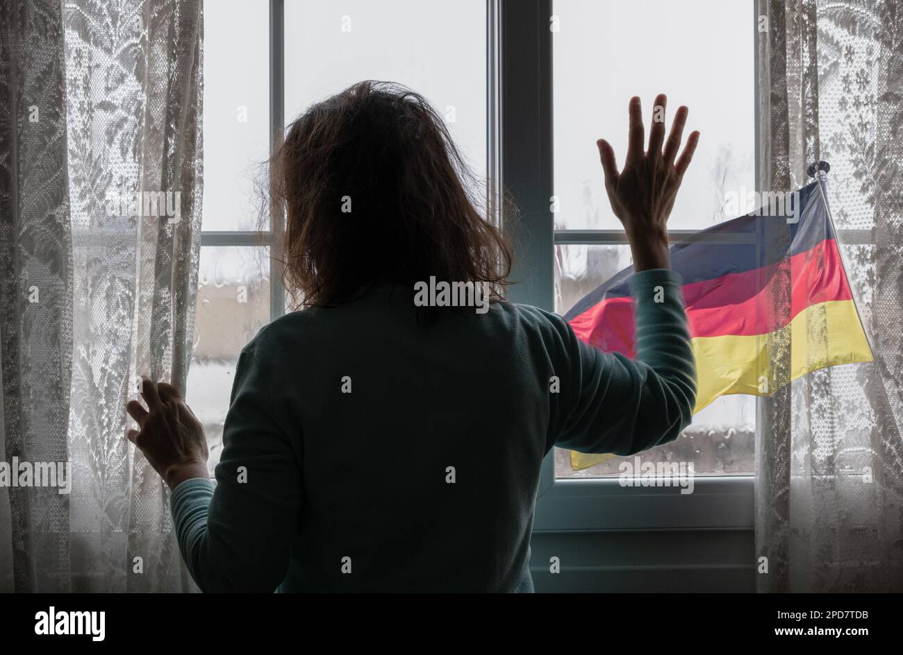 Eine Frau, die an Regentagen aus dem Fenster schaut. Flagge Deutschlands außerhalb. Konzept: Depression, häusliche Misshandlung, psychische Gesundheit, Menschenhandel, Sklaverei... Stockfoto