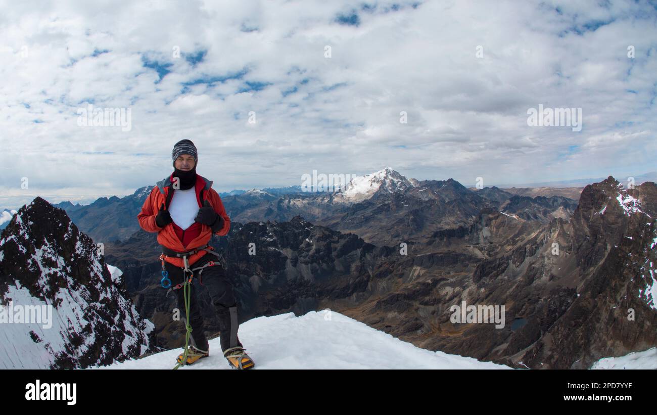 Junger, männlicher Kletterer, der an einem bewölkten Tag auf dem Gipfel des verschneiten Berges mit Helm, Stirnlampe, rotem Mantel und Seil steht Stockfoto