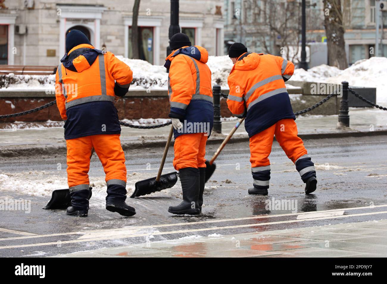 Drei Arbeiter säubern Schnee mit Schaufeln, Schneeräumen in der Stadt. Frühlingswetter, Straßenreinigung Stockfoto