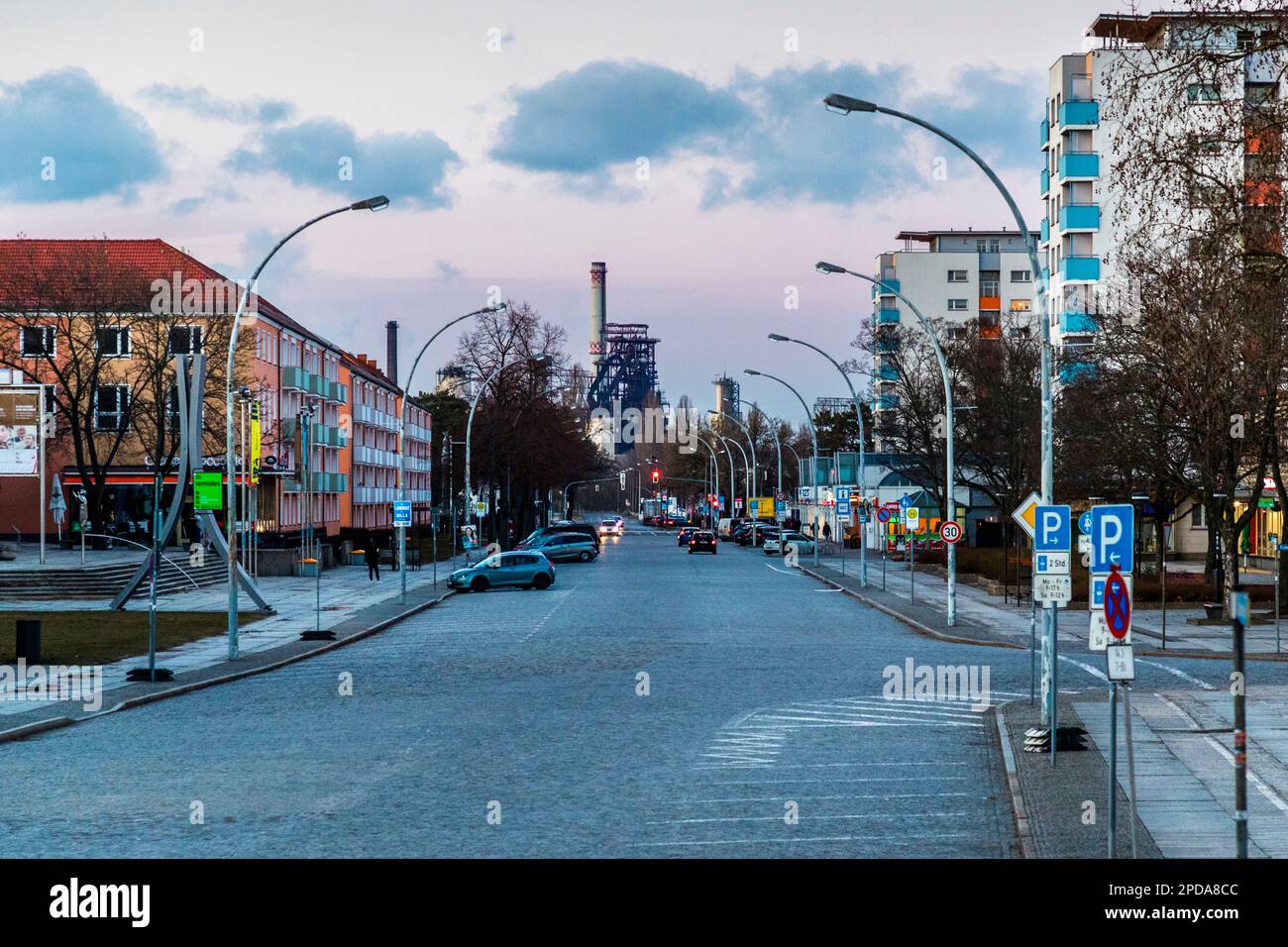 Mittlerer Abschnitt der Lindenallee (früher Leninallee) in Eisenhüttenstadt mit rhythmischen Abfolgen von Hochhäusern mit überstehenden Dächern und Geschäften. Am Ende der Straße sehen Sie den großen Hochofen. Das ehemalige EKO-Stahlwerk (Eisenhütten Kombinat Ost) von ArcelorMittal in Eisenhüttenstadt, Deutschland Stockfoto