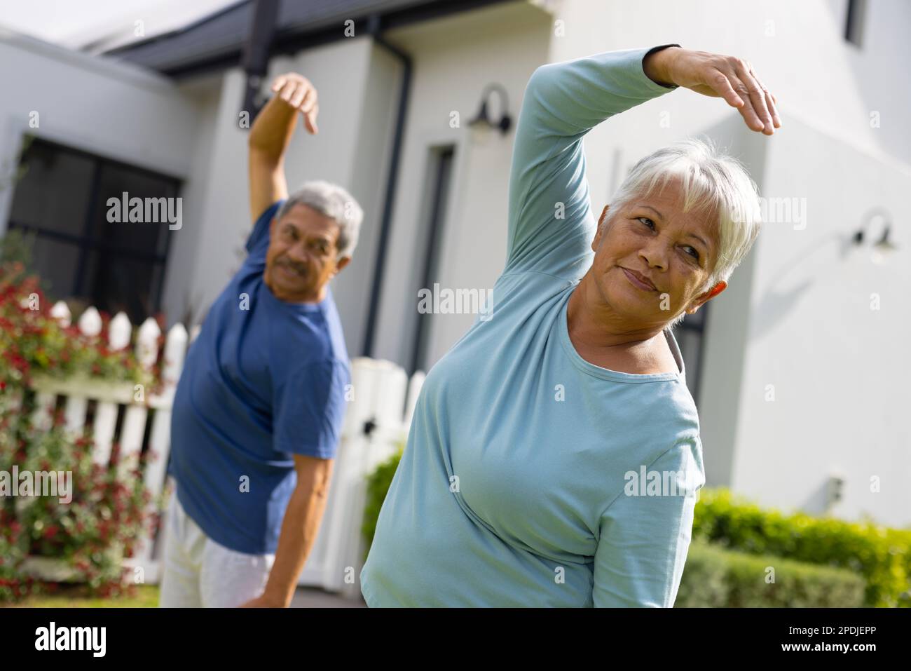 Ein zweirassistisches Seniorenpaar mit erhobenen Armen, das sich streckt, während er im Hof gegen das Haus steht Stockfoto