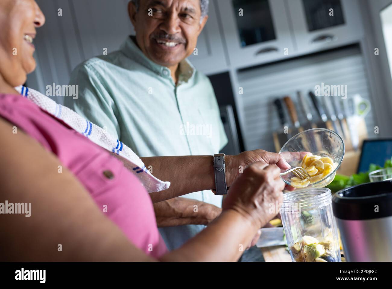 Ein lächelnder älterer Mann, der seine Frau ansieht, wie er Bananenscheiben mixt und Smoothie macht Stockfoto