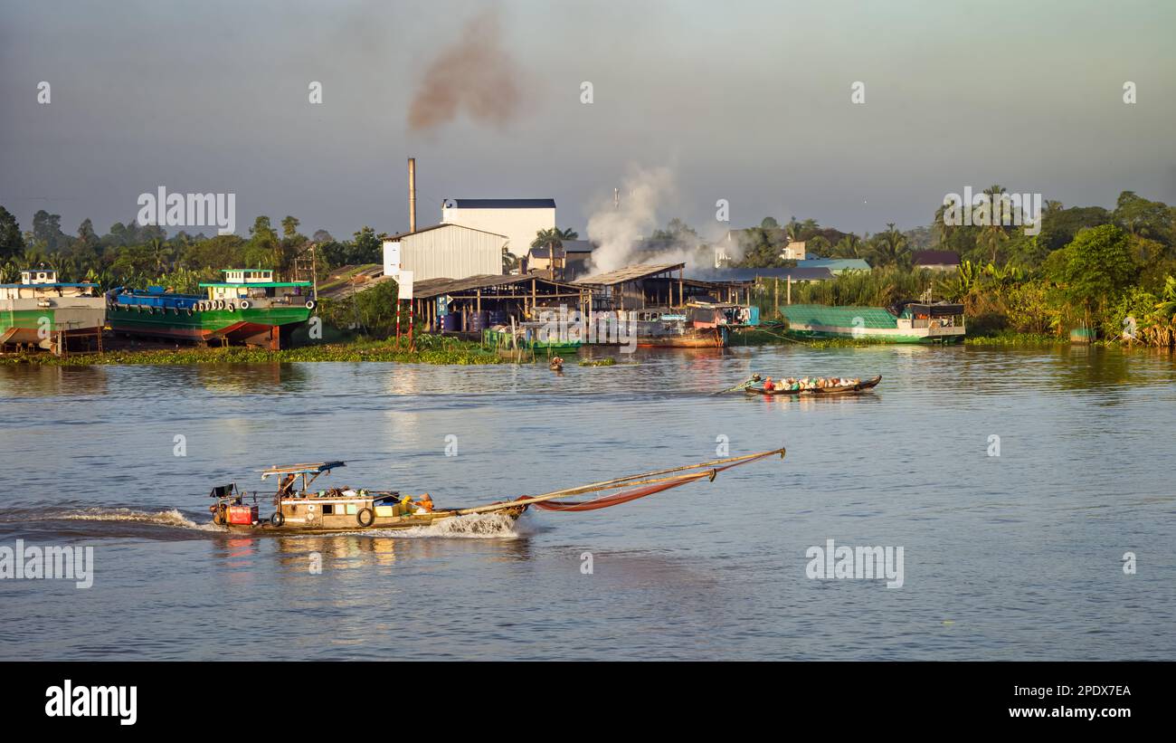 Ein hölzernes Fischerboot, bemannt von Mann und Frau, fährt an einer Fabrik vorbei, mit einem Boot, das Müll hinter sich transportiert, auf dem Mekong in der Nähe von Long Xuye Stockfoto