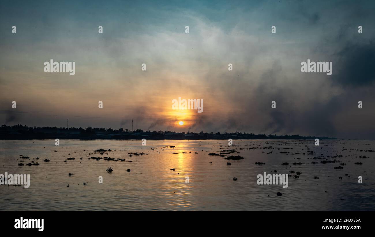 Rauchen aus Ziegelöfen, während die Sonne über dem Mekong-Fluss aufgeht, mit seinen Klumpen schwimmender Wasserhyazinthen am Long Xuyen im Mekong-Delta in Stockfoto