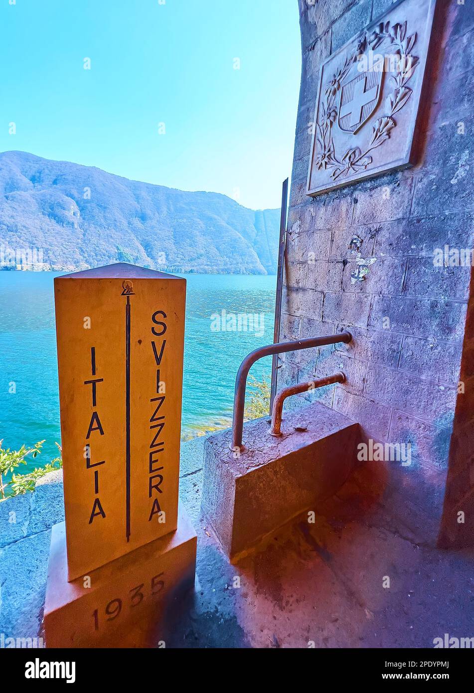 Der alte Grenzmast Italien-Schweiz am Ufer des Luganer Sees gegen die Lugano-Voralpen und die Tunnelwand mit Schweizer Wappen Stockfoto