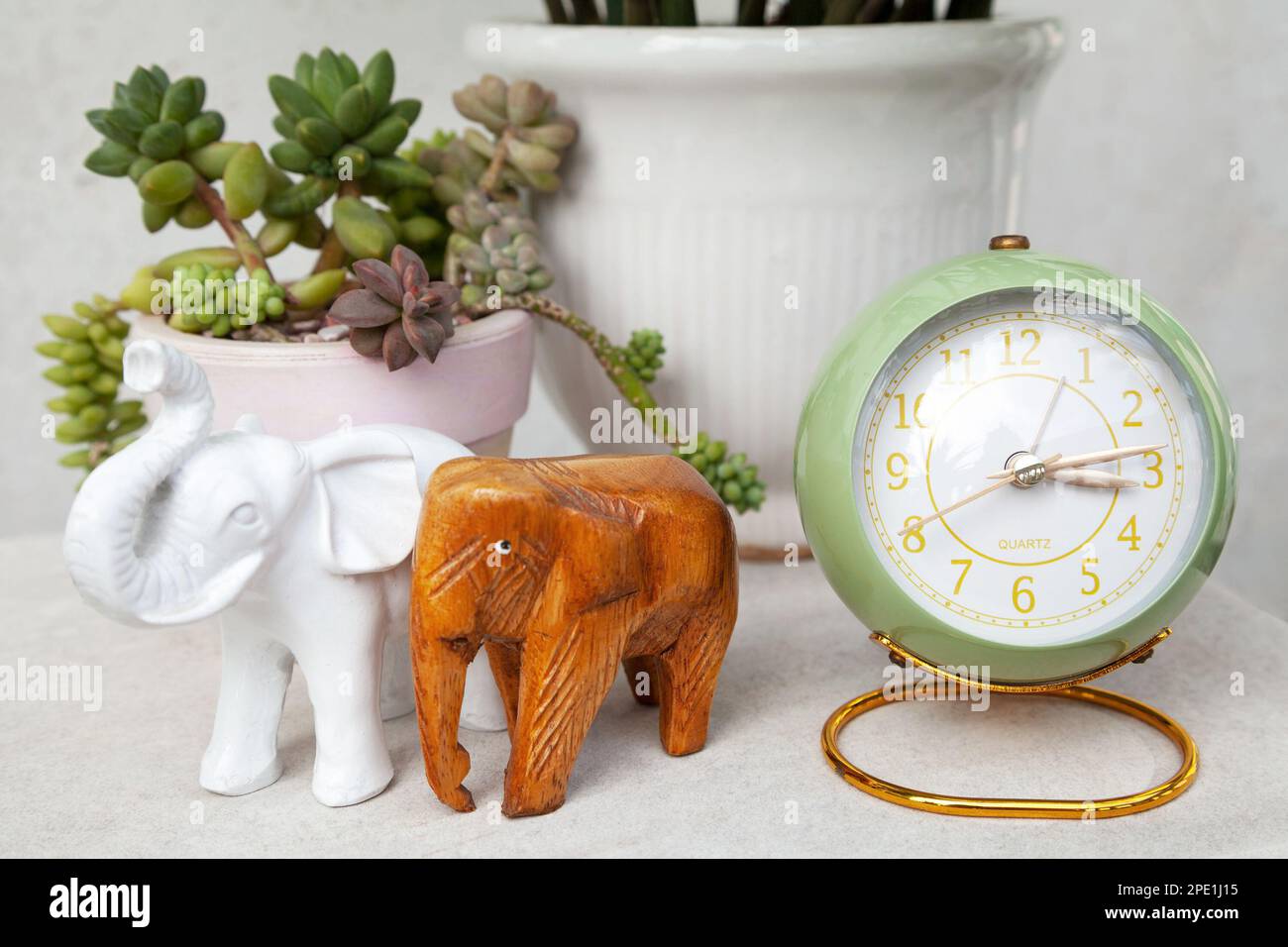Grüner Wecker im Vintage-Stil, der die Uhrzeit am Nachmittag anzeigt. Stilvolle rund um die Uhr, Elefantenfiguren und saftige Topfpflanzen auf einem Nachttisch Stockfoto